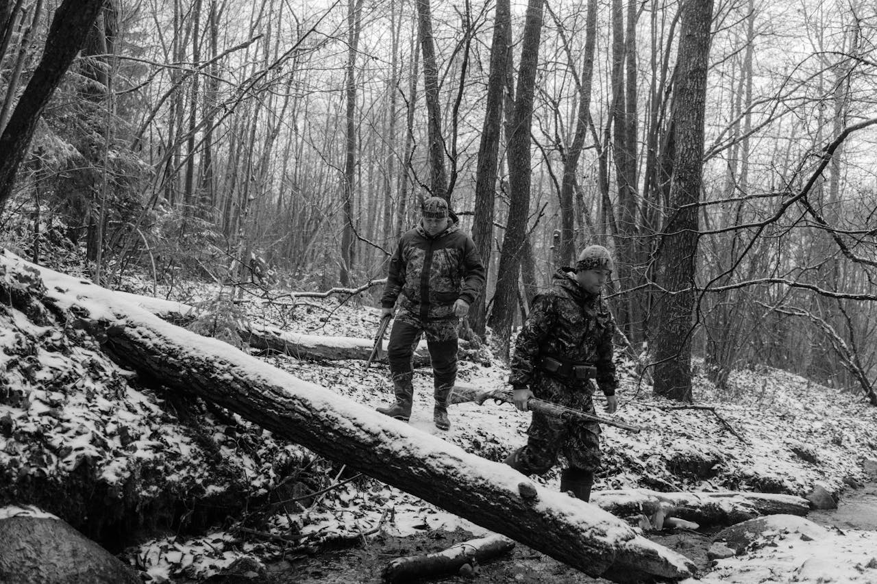 Home Two hunters in camouflage walking through a snowy forest, holding rifles. A monochrome winter scene.